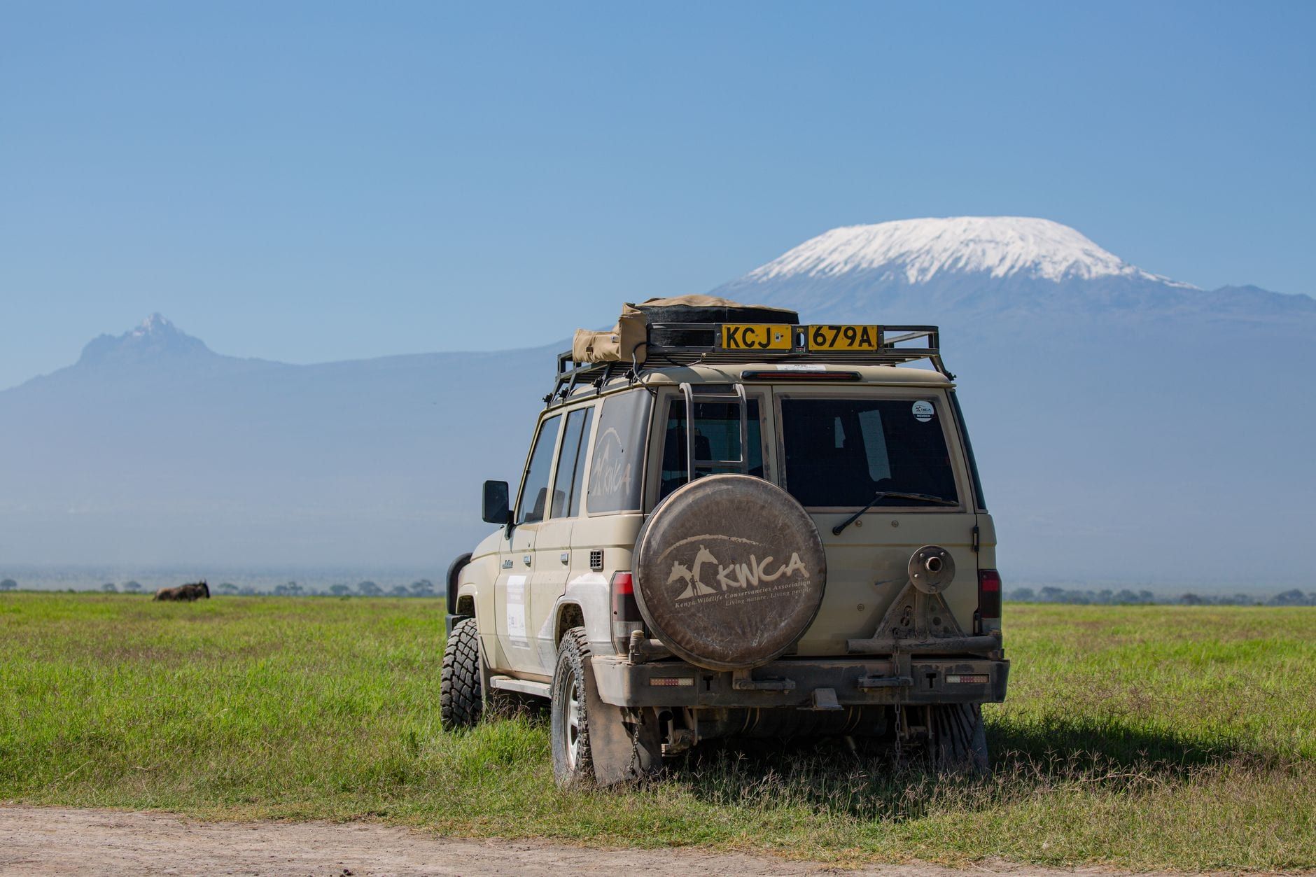 A safari vehicle parked on the grassy plains of Amboseli National Park with the snow-capped peak of Mount Kilimanjaro in the background.