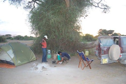 Campers set up a campsite with a pitched dome tent and vehicle beneath an acacia tree in Amboseli National Park.