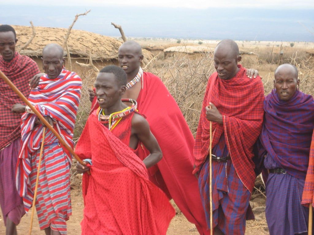 Maasai men dressed in traditional red shúkà garments dance in front of boma huts near Amboseli National Park.