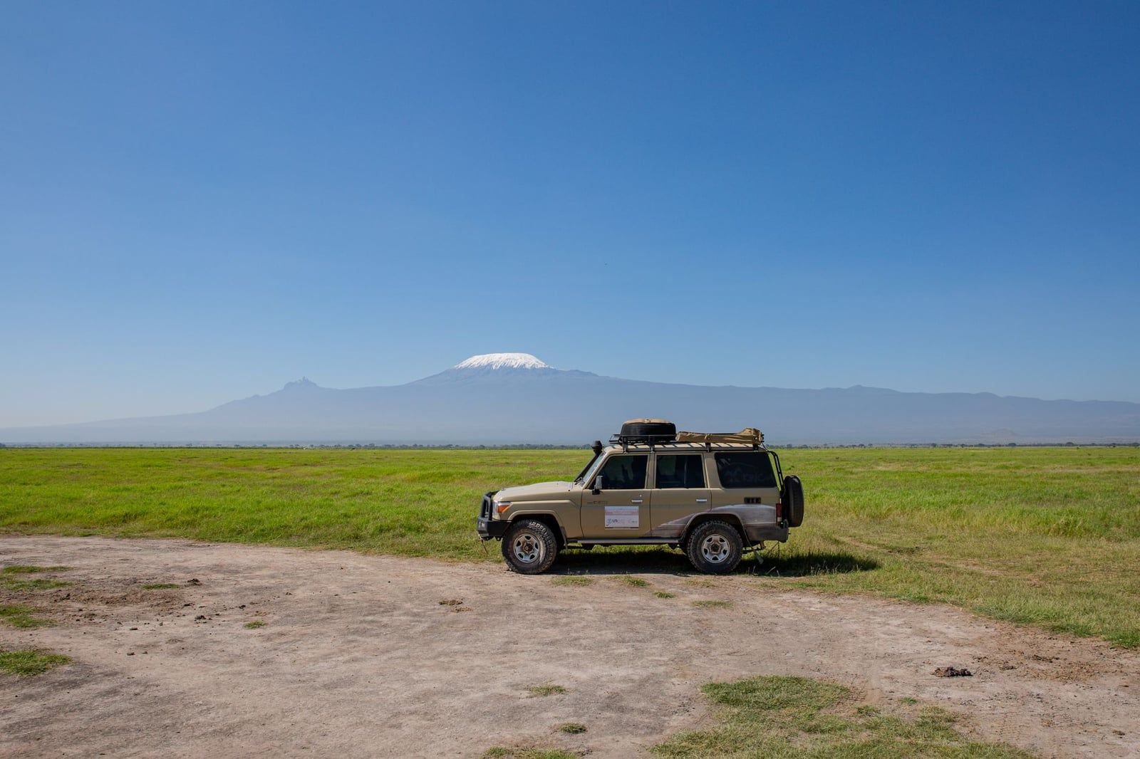 A safari 4x4 vehicle parked on a dirt track in Amboseli National Park with Mount Kilimanjaro visible across the plains.